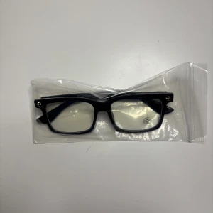 Svarta rektangulära glasögon med blommig detalj - Snygga svarta glasögon med rektangulär form och transparenta linser. Bågarna har en blommig detalj i silver på sidorna som ger en unik touch. Tillverkade i blank syntet, passar dig som gillar stilrena och moderna accessoarer.