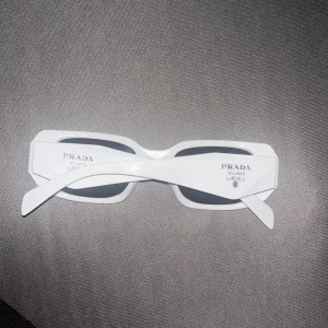 Vita rektangulära solglasögon från Prada - Säljer ett par snygga vita solglasögon från Prada med rektangulär form och svarta linser. Modellen har breda skalmar med Prada Milano-logga och coola geometriska detaljer. Perfekt för dig som vill sticka ut med en lyxig och trendig accessoar. Perfekt för alla väder