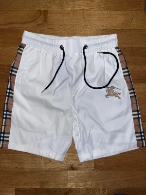 Vita Burberry shorts med rutiga sidor - Snygga vita shorts från Burberry med klassiska rutiga paneler på sidorna och guldfärgat ryttaremblem framtill. Elastisk midja med svart snörning och fickor på sidorna. Perfekta för sommaren och ger en clean, lyxig vibe.