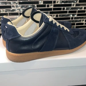 Maison Margiela sneakers blå - Maison Margiela sneakers i mörkblå mocka och skinn med kontrasterande vita snören och beige innersida. Snygg retrostil med gummisula och rund tå. Perfekt för dig som gillar stilrena och exklusiva sneakers med unik design. 