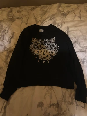 Svart Kenzo tröja med tigerbrodyr - Svart tröja från Kenzo med ikonisk tigerbrodyr och texten 'Kenzo Paris' i vitt framtill. Klassisk rund hals och långa ärmar. Tröjan är i mjuk bomull och har en normal passform som sitter snyggt på kroppen.