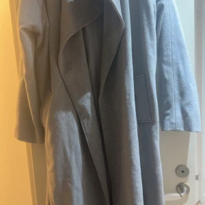Grå omlottkappa OC Trane XL - Säljer en stilren grå kappa från OC Trane i storlek XL. Kappan har omlottkrage, raka ärmar och stora fickor framtill. Perfekt för dig som gillar minimalistisk look och vill ha en snygg och enkel jacka till höst och vinter.