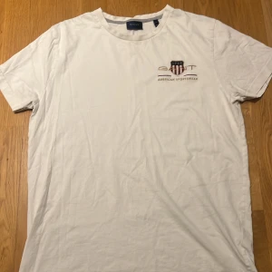 Vit t-shirt från GANT i bomull - Vit t-shirt från GANT med tryckt logga och emblem på bröstet. Klassisk rund hals och korta ärmar. Tillverkad i mjuk bomull som känns skön mot huden. Perfekt för dig som gillar stilren och enkel design med en sportig touch.