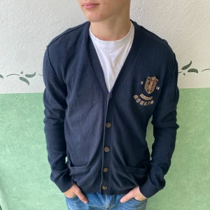 Mörkblå kofta från Morris, storlek L - Snygg mörkblå kofta från Morris med broderat emblem och text på bröstet. Koftan har v-ringad hals, knappar framtill och två fickor. Tillverkad i mjuk bomull, perfekt för lager-på-lager. Klassisk och stilren design med ribbade muddar.