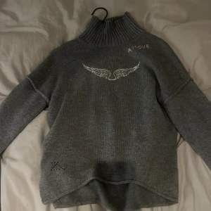 Grå polotröja Zadig & Voltaire - Grå stickad polotröja från Zadig & Voltaire med ribbad hög krage. Framsidan har ett vitt tryck med vingar och broderad text 'AMOUR' vid axeln. Tröjan har lång ärm och detaljerad hålstickning vid nederkanten. Perfekt för dig som gillar coola detaljer och fransk vibe.