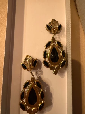 Stora svarta och guldiga statementörhängen (clips) - Säljer ett par maffiga statementörhängen i guldmetall med svarta stenar. Örhängena har droppformad design med flera svarta, slipade stenar i olika former och clip-on fäste. Perfekta för dig som vill sticka ut och addera lyxig känsla till din look.