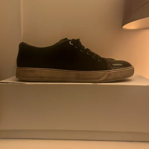 Lanvin mörkblå sneakers med lackad tå - Lanvin sneakers i mörkblått med snygg lackad tå och klassisk snörning. Skorna har en rund tå och platt sula i beige, med metallöglor och en stilren design. Materialet är en mix av skinn och lack, vilket ger en lyxig känsla. Perfekt för dig som gillar exklusiva sneakers.