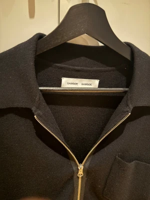 Svart kofta med dragkedja från Samsøe Samsøe - Svart tröja från Samsøe Samsøe med klassisk krage och hel dragkedja framtill. Tröjan har en bröstficka och är långärmad, tillverkad i mjukt stickat material som känns skönt mot huden. Perfekt för lager-på-lager och stilren look.