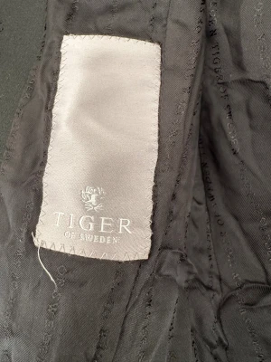 Svart kavaj från Tiger of Sweden - Klassisk svart kavaj från Tiger of Sweden med två knappar framtill, fickor med lock och snyggt figursydd passform. Fodrad insida med diskret mönster och märkets logga. Perfekt för en stilren look. storlek 38. 