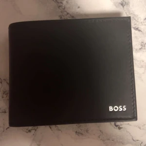 Svart plånbok från BOSS i skinn - Pris kan diskuteras. Kommer med låda Stilren svart plånbok från BOSS i äkta skinn med diskret logga i silver på framsidan. Plånboken har flera kortfack och ett myntfack, perfekt för att hålla ordning på dina kort och cash. Enkel och klassisk design som passar till allt. 