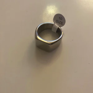 Silver Ring - Snygg silverfärgad ring i storlek 10 (2cm diameter, ~7.5cm omkrets) med fyrkantig platta framtill. Ringen har en modern och stilren design i blankt silvrigt metallmaterial. Perfekt för dig som gillar statement-smycken och vill ha något som sticker ut. PRISET KAN DISKUTERAS.