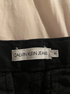 Svarta byxor från Calvin Klein Jeans - Snygga svarta byxor från Calvin Klein Jeans med diskret CK-logo vid fickan. Klassisk rak passform och midja med knapp och dragkedja. Materialet är mjukt och känns som bomull, perfekt för en clean och stilren look.