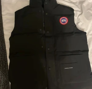 Svart dunväst från Canada Goose - Svart dunväst från Canada Goose med klassisk logotyp på bröstet och diskret broderad märkning på fickan. Västen har hög krage, dragkedja och tryckknappar framtill samt stora fickor. Materialet är polyester och bomull, perfekt för kyliga dagar.