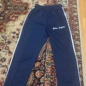 Palm Angels blå trackpants med vita ränder - Snygga marinblå trackpants från Palm Angels med vita sidoränder och logga tryckt på låret. Byxorna har elastisk midja och raka ben, perfekt för en sportig och trendig look. Materialet är mjukt och följsamt, vilket gör dem riktigt sköna att bära.