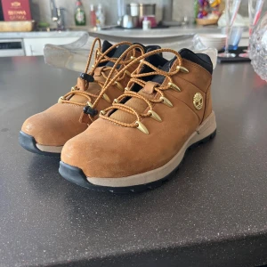 Bruna Timberland kängor med snörning - Säljer ett par snygga bruna Timberland kängor med robust svart sula och guldfärgade detaljer vid snörningen. Skorna har Timberland-logga på sidan och bak, samt svart vadderad kant upptill. Tillverkade i mocka med slitstark gummisula, perfekta för höst och vinter.