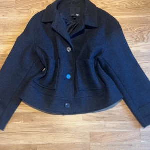 Mörkgrå/svart kappa  - Snygg mörkblå kavajjacka från 157 i storlek M. Jackan har klassisk krage, tre stora knappar framtill och två fickor. Materialet är ulligt och ger en stilren look. Passformen är lite loose och modellen är något croppad. Perfekt för dig som gillar enkel och clean stil.