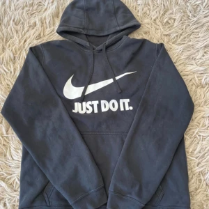 Svart Nike hoodie med tryck M - Svart hoodie från Nike i storlek M med klassiskt 'Just Do It.' tryck och swoosh-logga i vitt på bröstet. Tröjan har huva med snörning och en stor magficka. Materialet är 80% bomull och 20% polyester, vilket gör den mjuk och skön. Perfekt för chill eller träning. 