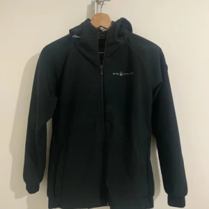 Svart vindjacka från Sail Racing - Svart vindjacka från Sail Racing med huva och hel dragkedja framtill. Jackan har diskret logga på bröstet och är designad med långa ärmar och praktiska fickor. Perfekt för dig som gillar stilren och sportig look.