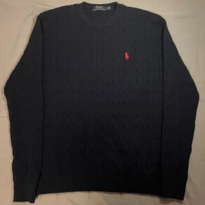 Mörkblå stickad tröja Polo Ralph Lauren - Snygg mörkblå stickad tröja från Polo Ralph Lauren med klassiskt kabelstickat mönster och rund hals. Tröjan har långärmad passform och en röd broderad logga på bröstet. Perfekt för dig som gillar stilrena och tidlösa plagg.