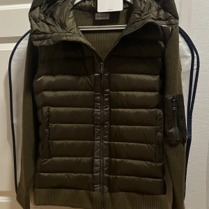 Moncler Cardigan  - Snygg olivgrön dunväst från Moncler med quiltad front och huva. Västen har stickade sidor och rygg i matchande färg, samt praktiska fickor med dragkedja och tryckknapp. Perfekt för lager-på-lager och har en modern, sportig look med detaljerad dragkedja.