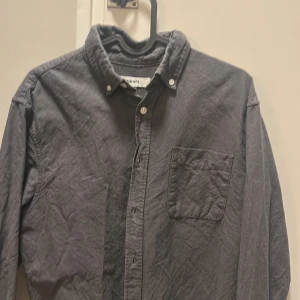 Mörkgrå skjorta från Kappahl - Snygg mörkgrå skjorta från Kappahl med klassisk krage och knappstängning framtill. Skjortan har en bröstficka och långa ärmar med manschettknappar. Tillverkad i mjuk bomull som känns skön mot huden. Perfekt för dig som gillar stilrena och enkla plagg.