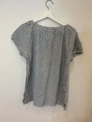 Randig blus med knyt i halsen - Säljer en luftig blus från Isolde med svartvita ränder. Blusen har korta ärmar, rundad halsringning med knyt och är tillverkad i ett lätt bomullstyg. Perfekt för dig som gillar en avslappnad och trendig stil.