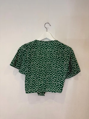 Grön blommig knytblus från SHEIN - Grön blommig blus från SHEIN med vita små blommor, kort ärm och croppad passform. Toppen har v-ringning och knyt framtill för en söt och trendig look. Perfekt för dig som gillar färg och mönster.