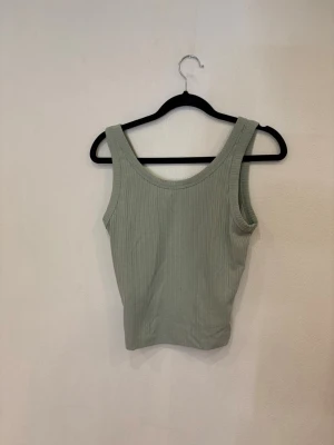 Ljusgrön ribbad linnetopp - Säljer en ljusgrön ribbad linnetopp med breda axelband och rundad halsringning. Toppen är ärmlös och har en enkel, stilren design som passar perfekt till jeans eller kjol. Materialet är mjukt och stretchigt för en bekväm passform.