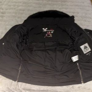 Svart Moose Knuckles dunjacka med päls - Säljer en svart dunjacka från Moose Knuckles med fet päls på kragen och snygga metalldetaljer. Jackan har broderad logga och motiv på insidan, dragkedjor på ärmar och bröst, samt knappar framtill. Perfekt för dig som vill ha en varm och stilren jacka med attityd.