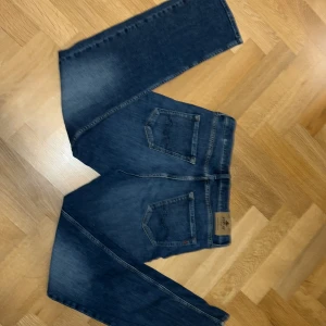 Blå jeans Replay Thad 16A - Snygga blå jeans från Replay, modell Thad, med klassisk femficksdesign och tydliga slitningar på benen. Jeansen har raka ben och normal passform, med Replay-logga på bakfickan och läderpatch i midjan. Perfekt för dig som gillar stilrena och tidlösa jeans.