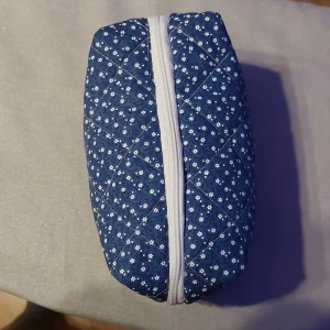  Necessär - En necessär i blått tyg med små vita blommor och vita sömmar i rutmönster. Necessären har en vit dragkedja upptill och är perfekt för smink, pennor mm. Rektangulär form och mjuk känsla, passar lätt i väskan eller på resan. Bredd: ca 9cm Längd: 19cm djup: 10cm 