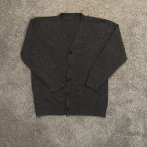 Mörkgrå kofta med knappar - Mysig mörkgrå kofta med v-ringad hals och fyra svarta knappar framtill. Tillverkad i mjukt stickat material som känns skönt mot huden. Perfekt att slänga på sig när det är lite kyligt ute. Långa ärmar och ribbade muddar vid ärmslut och nederkant.    Passar om man är inför för 165cm