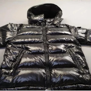 Svart pufferjacka från Moncler - Säljer en svart, glansig pufferjacka från Moncler med ikonisk logga på ärmen och coola detaljer som grov dragkedja och serietidningsmärke på insidan. Jackan har hög krage, två sidofickor med dragkedja och ett stilrent, quiltat mönster. Perfekt för dig som vill ha en snygg vinterlook.