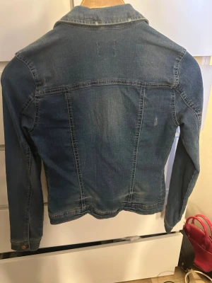 Blå jeansjacka från ONLY, strl 34 - Snygg klassisk jeansjacka från ONLY i blå denim. Jackan har långa ärmar, två bröstfickor med knapp och metallknappar framtill. Perfekt för dig som gillar en tidlös och chill stil. Passar dig som vill ha en jacka med normal passform och coola detaljer.