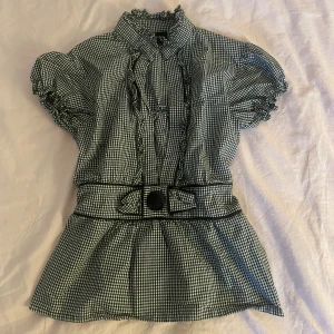 Rutig blus  - Blus med svartvitt rutigt mönster, puffärmar och volangdetaljer vid krage och ärmslut. Blus har markerad midja med dekorativt bälte och stor knapp framtill. Strl S