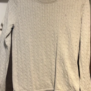 grå tröja med kabelmönster - Snygg ljusbeige stickad tröja med klassiskt kabelmönster. Tröjan har rund hals och långa ärmar, perfekt för dig som gillar en enkel men stilren look. Passar dig som vill ha en mjuk och bekväm tröja till hösten eller vintern.