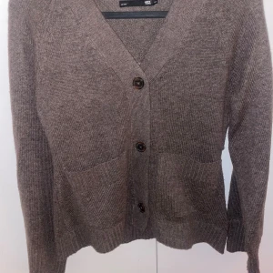 Brun stickad kofta från lager157  - Mysig brun stickad kofta från 157 i storlek XS. Koftan har två stora fickor framtill, v-ringad hals och stängs med knappar. Perfekt för lager-på-lager och chill dagar. Ribbstickade muddar vid ärmslut och nederkant ger extra komfort.