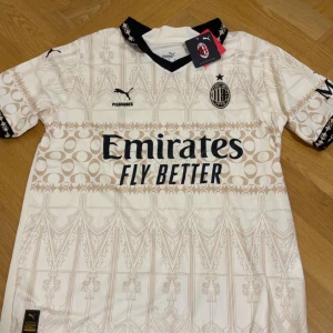 Begie Ac Milan tröja  - AC Milan fotbollströja i beige med unikt grafiskt mönster och svarta detaljer.  svart V-ringad krage och klubbmärke på bröstet. Materialet är lätt och andas, perfekt för match eller träning. Den är i storlek S men kan även passa M. Priset kan diskuteras!