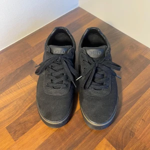 Svarta sneakers från Polo Ralph Lauren - Snygga svarta sneakers från Polo Ralph Lauren med klassisk design. Skorna har ovandel i textil och detaljer i skinn, rund tå och snörning framtill. Diskret Polo-logga på sidan och bak på hälen. Perfekta för en clean och stilren look.