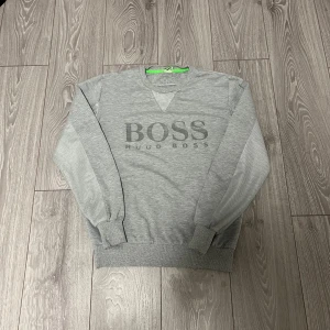 Grå sweatshirt från Hugo Boss - Snygg grå sweatshirt från Hugo Boss med stor BOSS-logga framtill. Tröjan har rund halsringning, ribbade muddar och långärmad design. Perfekt för en clean och stilren look. Materialet känns mjukt och bekvämt mot huden.