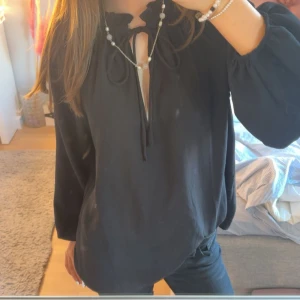Svart blus med knyt och volang - Säljer en svart blus med långa ärmar och volangdetalj vid halsen. Blusen har knytning framtill och en lätt v-ringning. Materialet känns mjukt och luftigt, perfekt för en chill och stilren look.