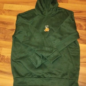 Grön hoodie med nallebrodyr - Mörkgrön hoodie med huva och lång ärm. På bröstet finns en broderad nalle i beige och vitt som ger en söt och lekfull touch. Tröjan är i mjukt material och har klassisk passform med ribbade muddar vid ärmslut och nederkant.