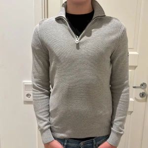Grå half zip tröja från Pelle P - Snygg grå tröja från Pelle P med half zip och hög krage. Tröjan är ribbstickad och har lång ärm samt logga på ärmen. Perfekt för lager-på-lager och chill stil. Passar dig som gillar klassisk men sportig look. Nypris 1500