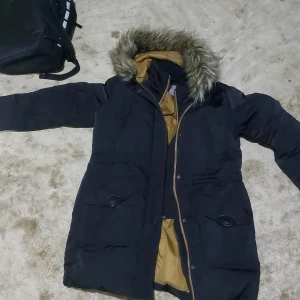 Svart parkas med fuskpäls från Studio Moda - Säljer en svart parkas från Studio Moda med varm fuskpäls runt huvan och snyggt bronsfärgat innerfoder. Jackan har stora fickor framtill, dragkedja och knappar. Perfekt för kalla dagar och har en klassisk, längre modell.