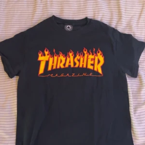 Svart Thrasher t-shirt med flames - Svart t-shirt från Thrasher med ikoniskt flames-tryck i gult och orange på bröstet. Klassisk passform och rund hals. Perfekt för dig som gillar streetwear och skatekultur. Materialet är mjuk bomull.