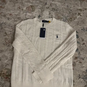 Vit kabelstickad tröja Polo Ralph Lauren - Vit kabelstickad tröja från Polo Ralph Lauren med klassisk rund halsringning och broderad marinblå logga på bröstet. Tröjan har ribbade muddar och är långärmad, perfekt för lager-på-lager under kyliga dagar.