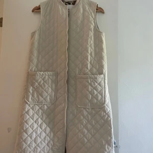 Lång quiltad beige väst med leopardfoder - Lång, ärmlös quiltad väst i beige med stora fickor framtill och dragkedja. Insidan har ett coolt leopardmönstrat foder i bruna och svarta toner. Perfekt för lager-på-lager och ger en trendig touch till din outfit.