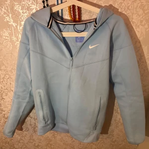 Nike NOCTA ljusblå hoodiejacka S - Nike NOCTA hoodiejacka i ljusblått med dragkedja framtill, huva och två fickor med dragkedja. Jackan har NOCTA-logga på ärmen och bröstet samt vita detaljer vid kragen. Perfekt för dig som gillar streetwear och sportig stil. Använd ett fåtal gånger 