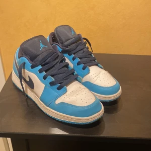Nike Air Jordan 1 Low blå/vit - Säljer ett par Nike Air Jordan 1 Low i blått och vitt med mörkblå detaljer och snörning. Skorna har klassisk Jordan-logga på hälen och tungan, samt vit platt sula. Perfekta för dig som gillar streetwear och vill sticka ut med snygga sneakers. Skorna är cresade fram över tårna. Priset kan diskuteras.