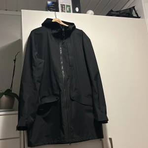 Svart regnjacka från McKinley XXL - Svart regnjacka från McKinley i storlek XXL. Jackan har huva, dragkedja framtill och två stora fickor med lock. Tillverkad i syntetmaterial som står emot regn och vind, perfekt för blöta dagar. Enkel och stilren design med logga på ryggen. knappt använd